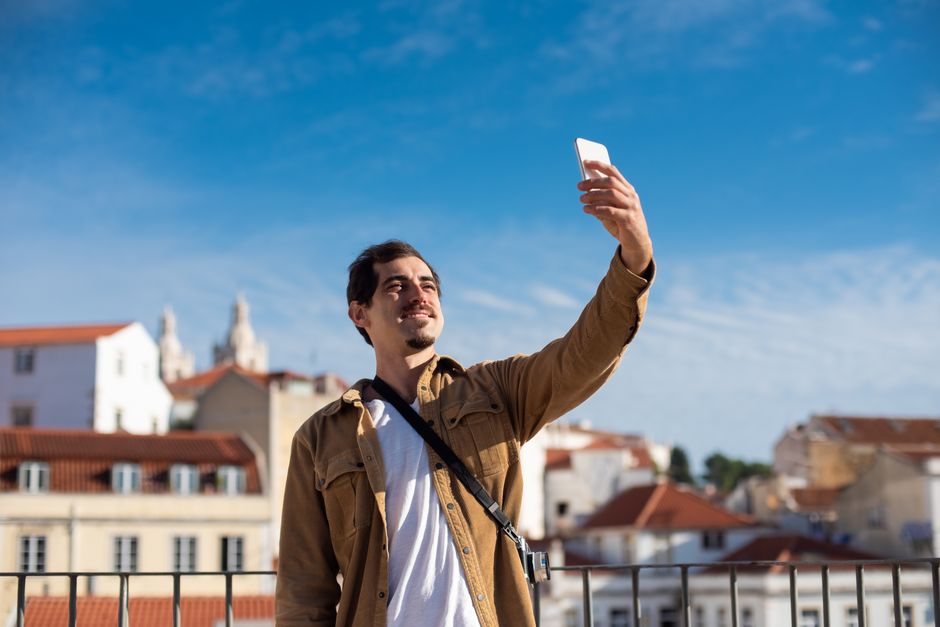 A man capturing a selfie against a picturesque sunlit cityscape with a clear blue sky.
