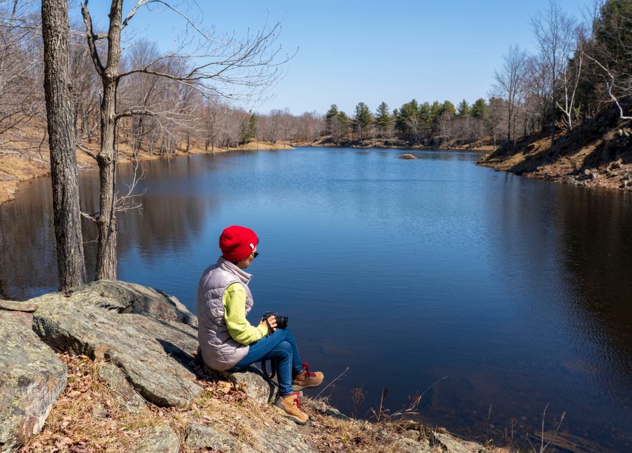 A woman in a red hat relaxes on rocks beside a tranquil lake on a crisp fall day.