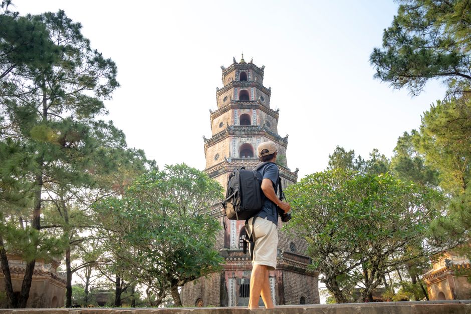 Side view of anonymous male tourist with backpack standing near green trees and old tower of temple during trip in Vietnam