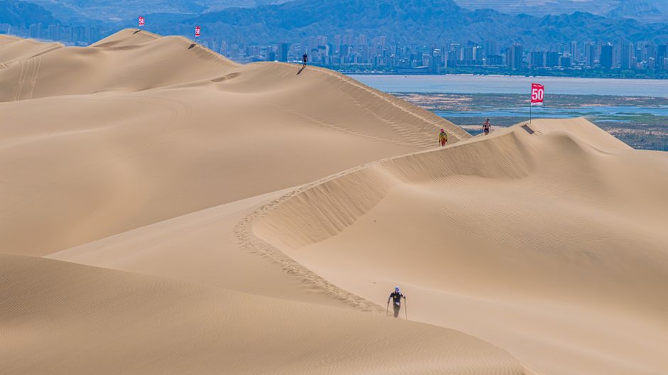 Hikers traverse vast desert dunes with a city skyline in the background, showcasing adventure and contrast.