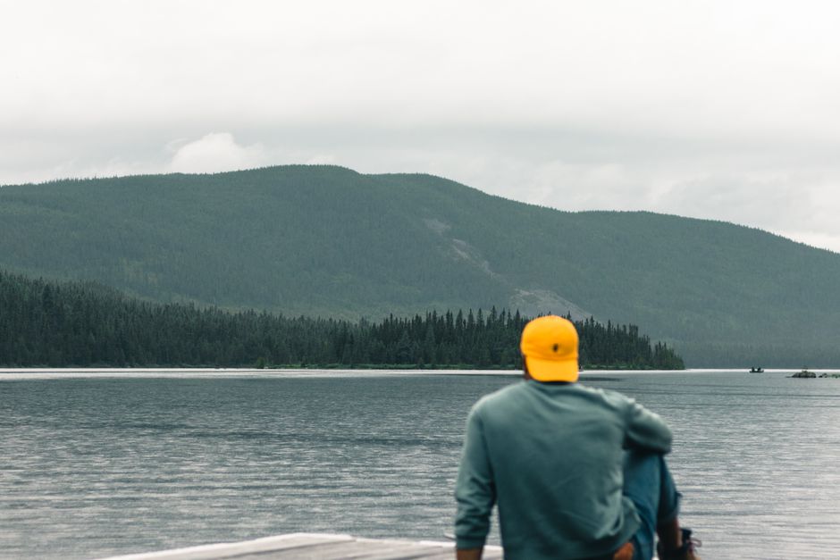 Man in yellow cap sits by a tranquil lake with mountain backdrop under overcast sky.