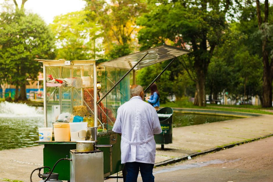An elderly vendor stands behind a street food cart in a lush urban park, serving customers.