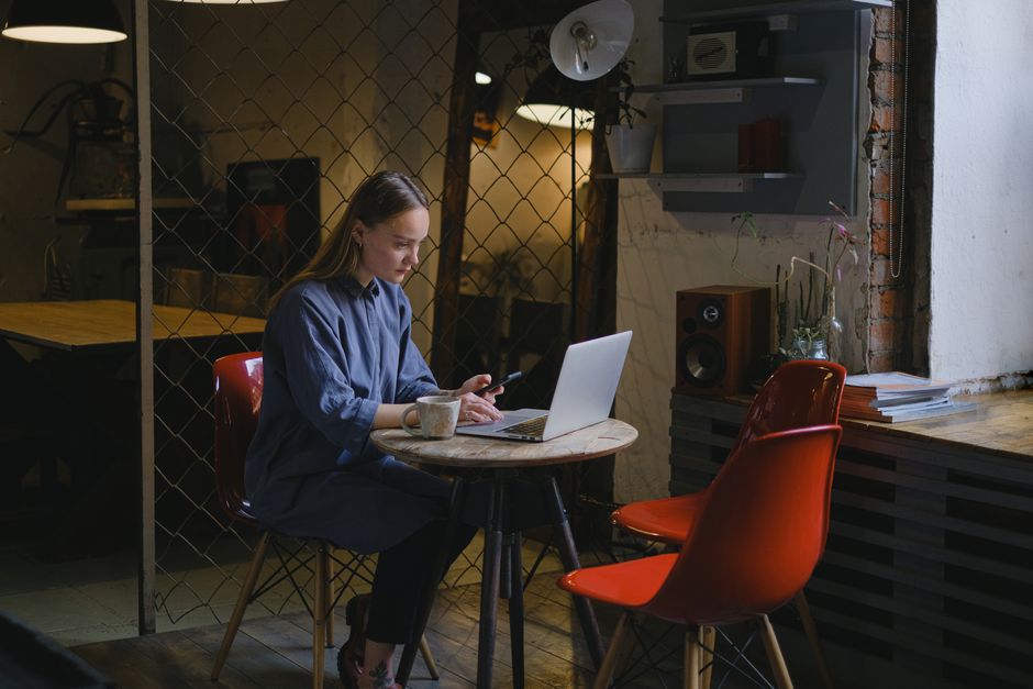 Side view of concentrated female freelancer with smartphone in hand surfing netbook while sitting at table with cup of coffee in cafeteria