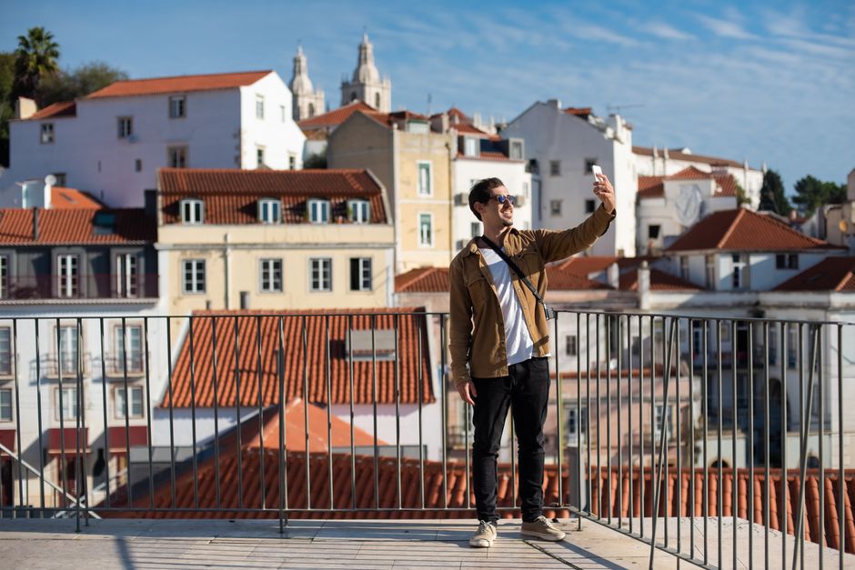 Man in sunglasses smiles while taking a selfie in Alfama, Lisbon with iconic rooftops in the background.