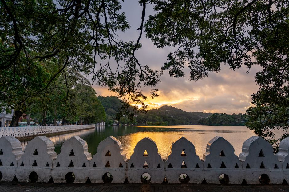 A picturesque view of a sunset over Kandy Lake with lush greenery and traditional architecture in the background.