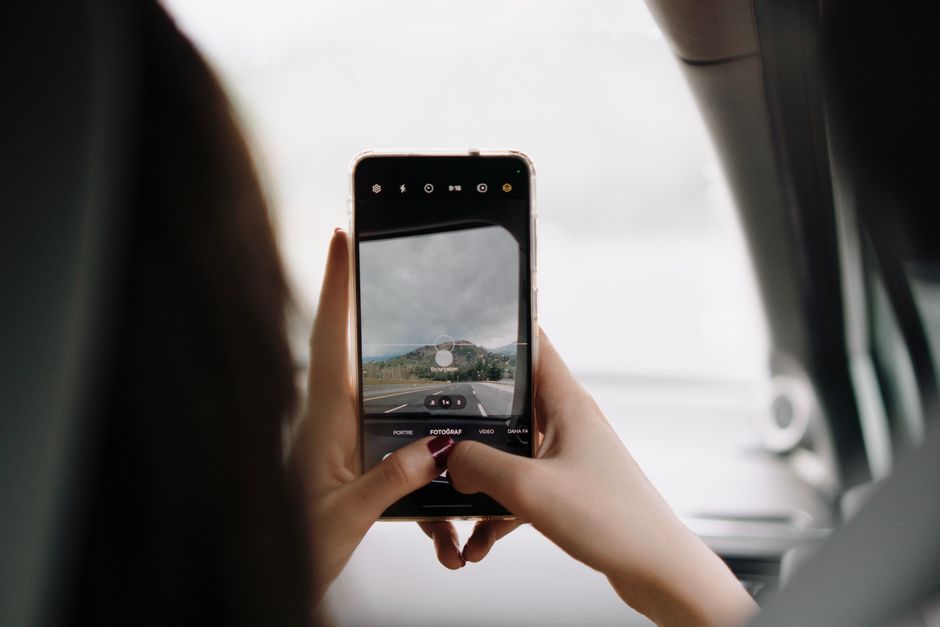 A person using a smartphone to capture a scenic view through the car window during a day trip.