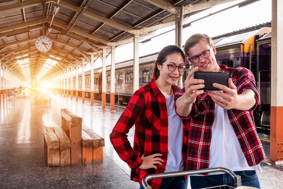 A smiling couple takes a selfie at an urban train station, showcasing travel and fashion.